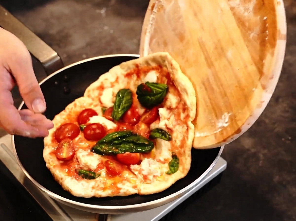 Una pizza transfiriéndose desde una pala de madera a una sartén mientras un soplete está listo cerca.