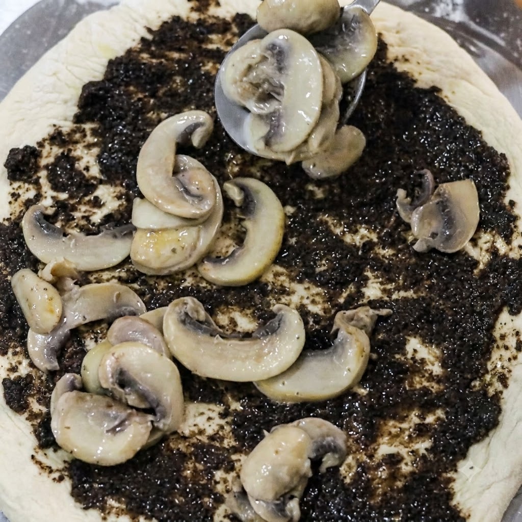 Cuchara colocando rodajas de champiñones cocinados sobre una base de pizza cubierta con pasta de trufa negra.