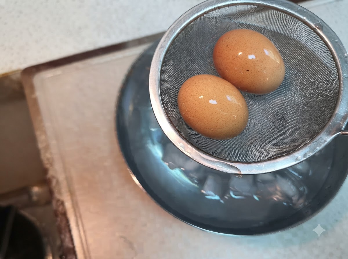 Dos huevos hervidos reposando dentro de un colador de malla metálica sostenido sobre un tazón de agua con hielo.