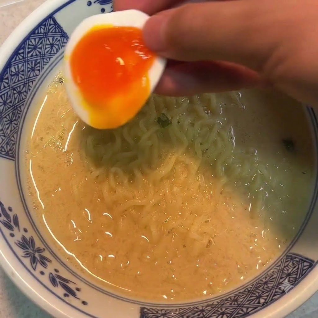 Una mano sosteniendo la mitad de un huevo pasado por agua sobre un tazón de fideos.