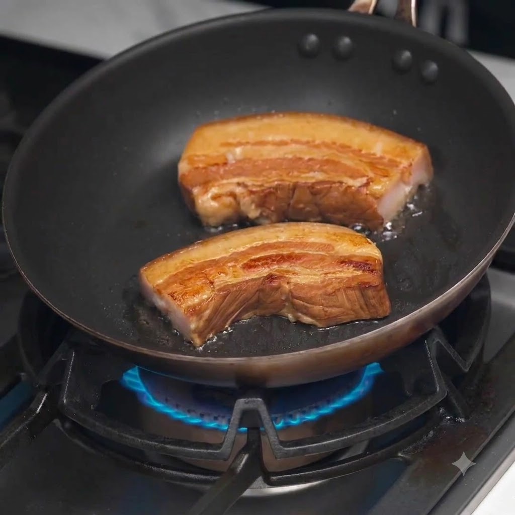 Rodajas gruesas de panceta de cerdo estofada dorándose en una sartén negra caliente sobre una estufa de gas.