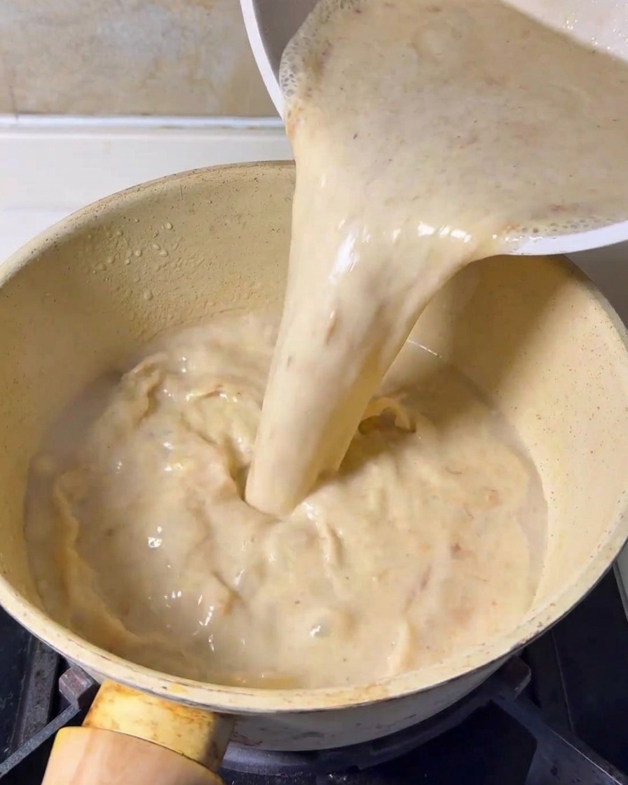 Base de sopa cremosa y licuada siendo vertida desde un recipiente en una olla de cocina amarilla sobre una estufa.