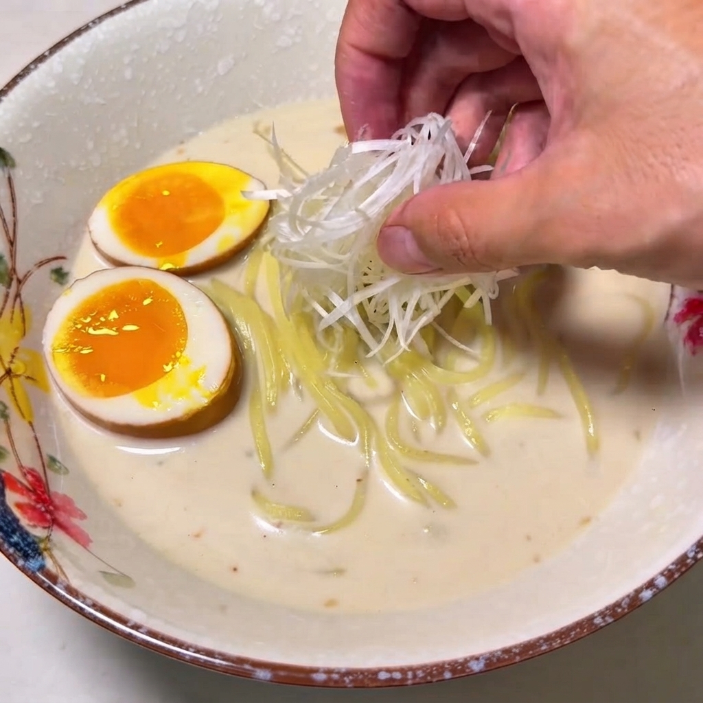 Una mano colocando un montón de cebollinas blancas trituradas en un tazón de ramen.