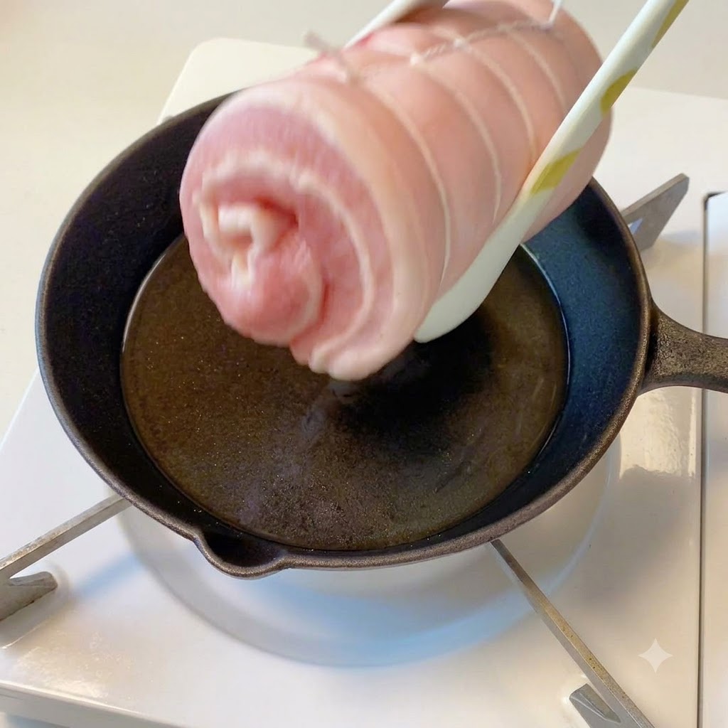 El cilindro de cerdo atado sostenido con pinzas sobre una sartén caliente con aceite.