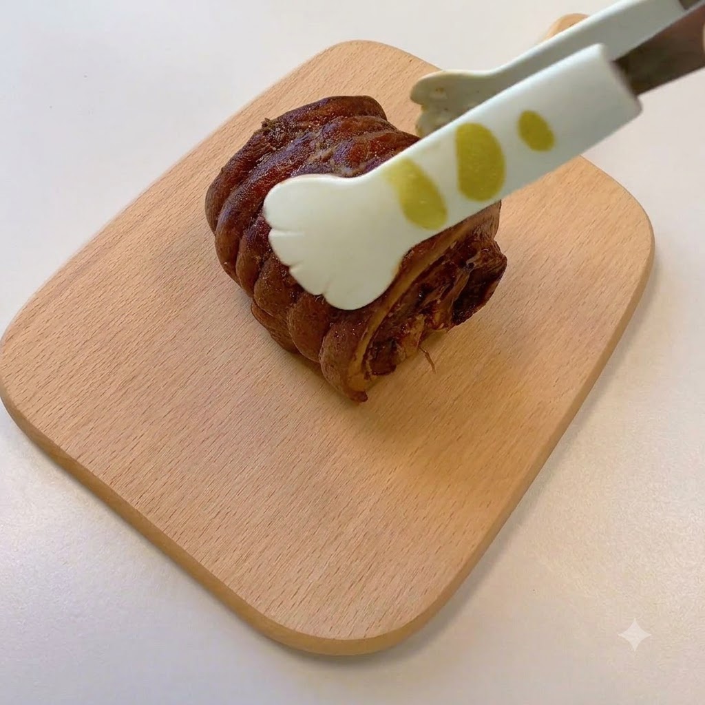 Unas pinzas sosteniendo una pieza de cerdo chashu enrollada y cocida reposando sobre una tabla de cortar de madera.