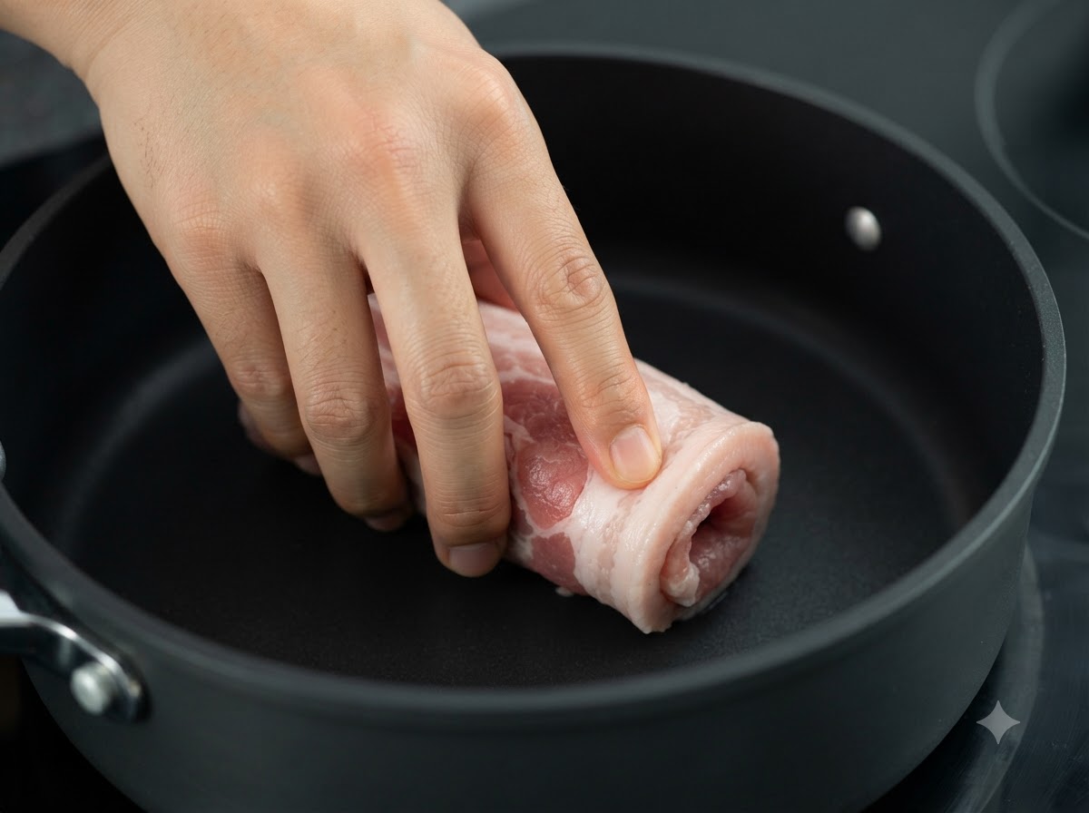 El rollo de panceta de cerdo atado dorándose en una sartén oscura a fuego.