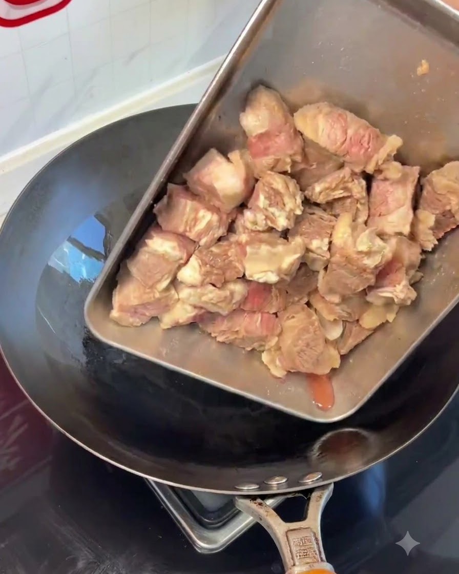Trozos de costilla de res blanqueados siendo vertidos desde una bandeja de acero a un wok caliente.