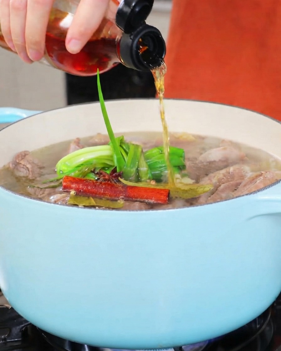 Vino de cocina siendo vertido desde una botella pequeña en una olla de estofado de color claro.
