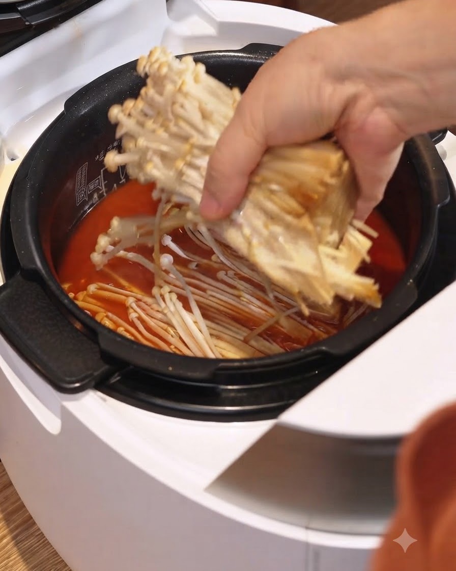 Un manojo de hongos enoki colocado en el caldo de estofado rojo dentro de una olla arrocera eléctrica blanca.