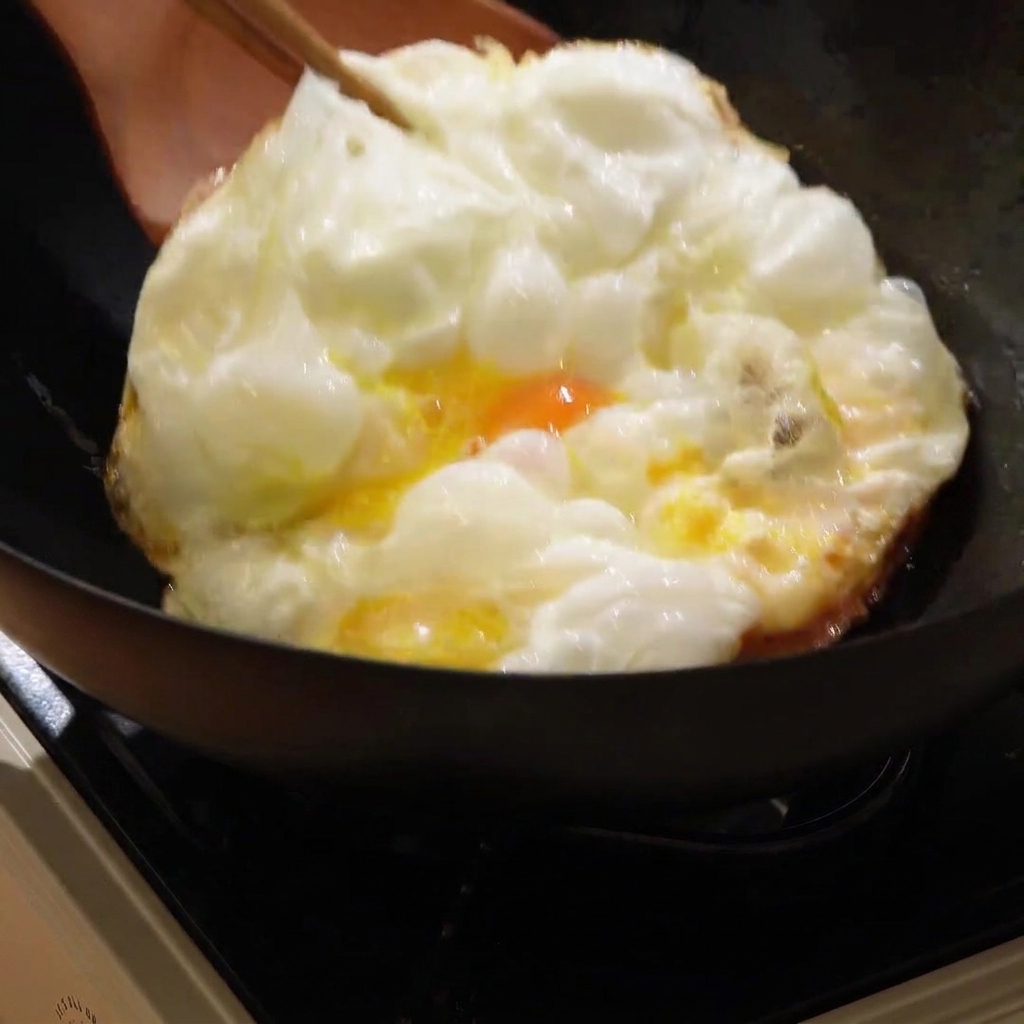 Un huevo frito grande de color marrón dorado siendo volteado en un wok caliente con una espátula de madera.