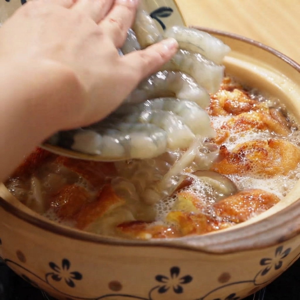 Camarones crudos pelados siendo añadidos a una olla de barro burbujeante llena de sopa y trozos de huevo frito.