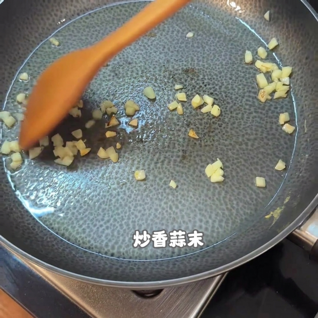 Ajo picado y trozos de comida siendo revueltos con una espátula de madera naranja en un sartén antiadherente.