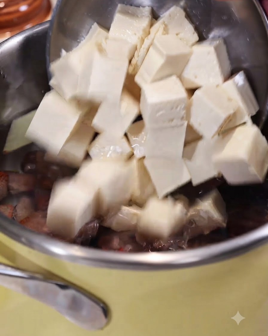 Tofu en cubos deslizándose desde una cuchara ranurada hacia una olla con agua hirviendo, jengibre y almejas.