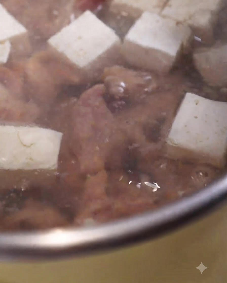Rodajas de cerdo marinadas cocinándose en una olla de sopa burbujeante junto con cubos de tofu blanco.