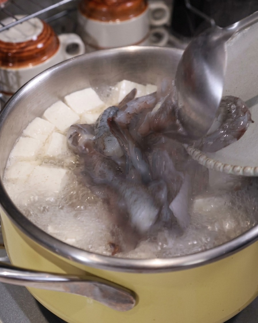 Un cucharón añadiendo trozos de calamar crudo y camarones a una olla hirviendo de sopa con tofu y cerdo.