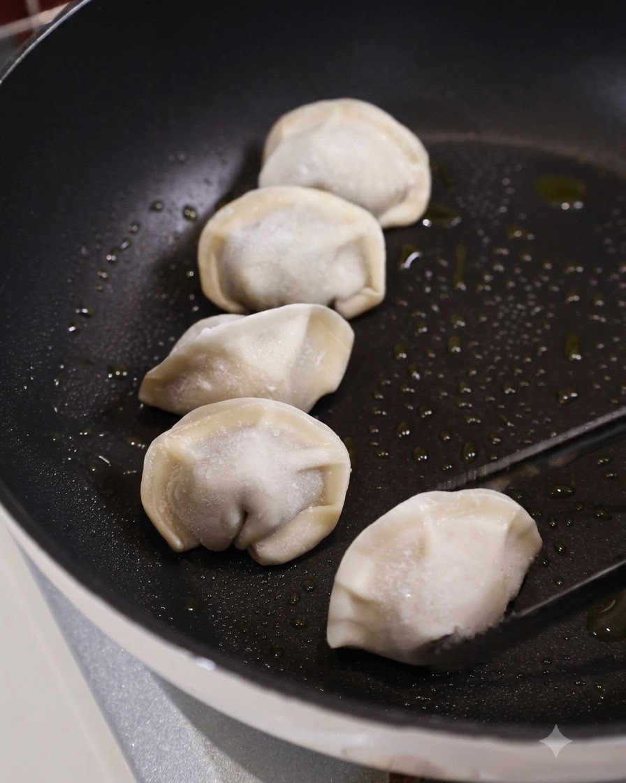 Pinzas de cocina colocando un dumpling congelado crudo en una sartén plana oscura y aceitada junto a varios dumplings más.