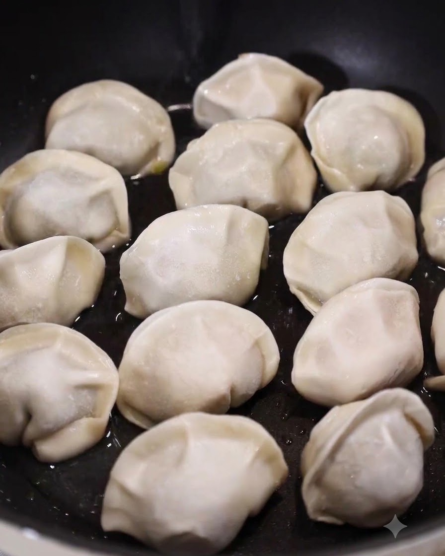 Una sartén plana llena de dumplings crudos reposando en una capa poco profunda de agua burbujeante y aceite.