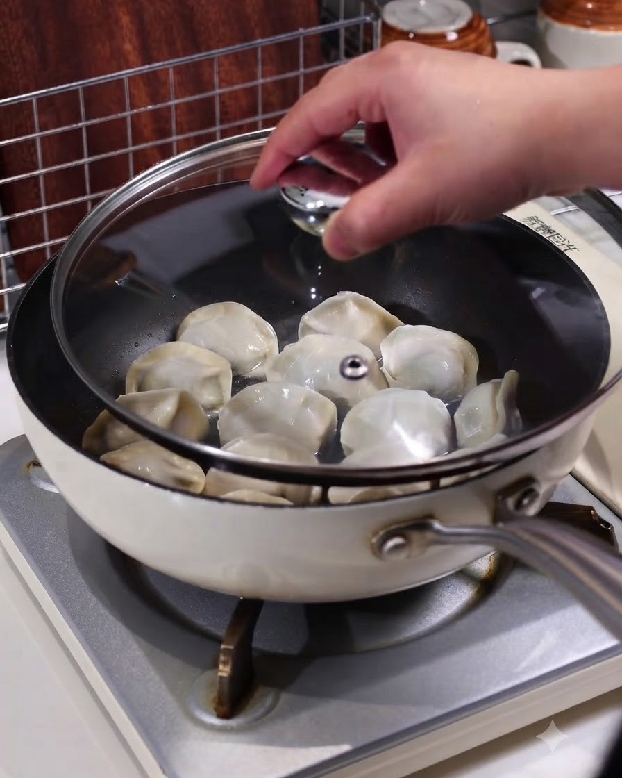 Una mano colocando una tapa de vidrio transparente en una sartén con dumplings al vapor en una estufa.