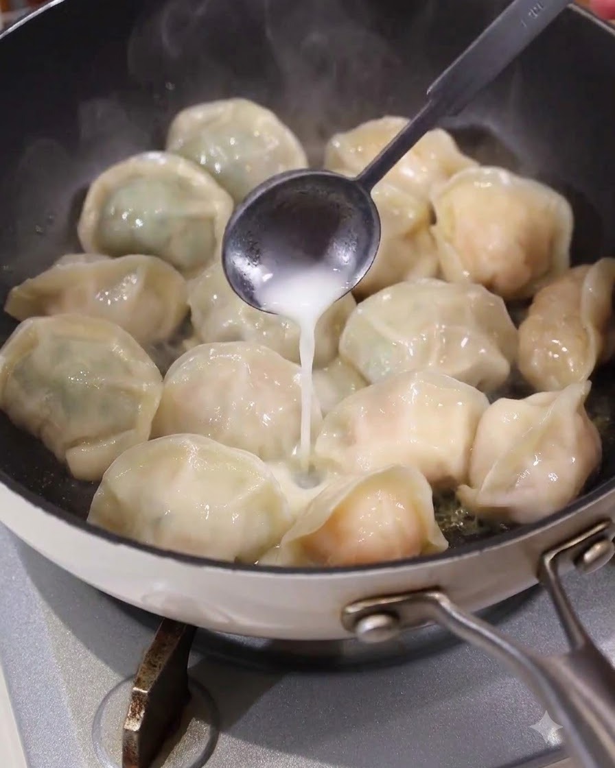 Una cuchara vertiendo una pasta líquida blanca de manera uniforme en los espacios entre los dumplings en una sartén caliente.