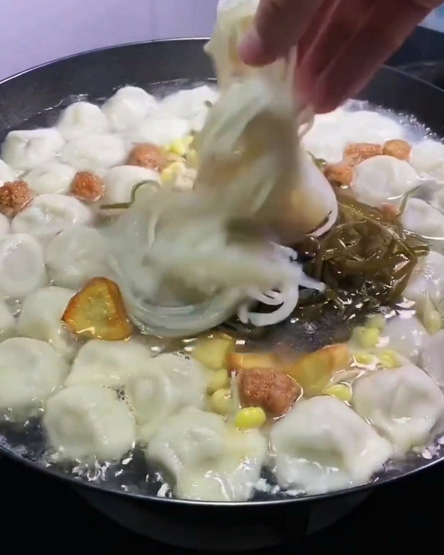 Una mano colocando alga kombu triturada y fideos de celofán translúcidos en una olla de dumplings hirviendo con caldo.