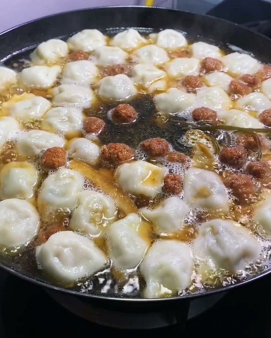 Salsa de soja oscura siendo añadida a una olla grande de dumplings hirviendo e ingredientes, aportando un color intenso.