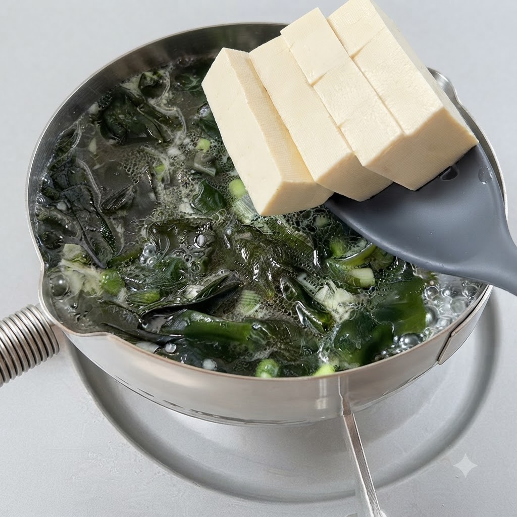 Cubos de tofu siendo transferidos a una olla burbujeante de sopa de algas con una espumadera.