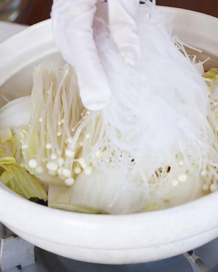Fideos de cristal remojados siendo colocados sobre la capa de hongos enoki y repollo.