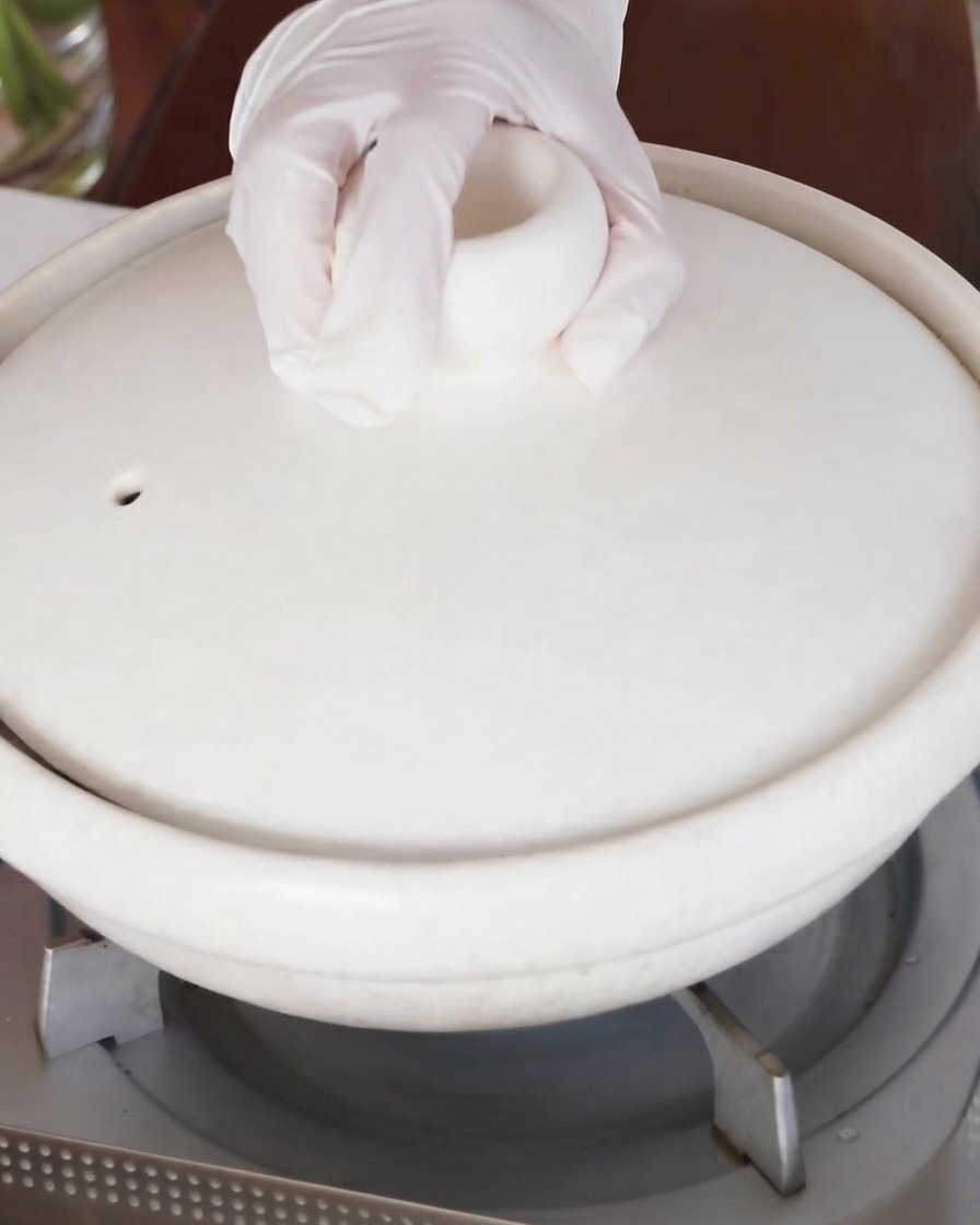Una mano colocando la tapa en la olla de estofado de cerámica blanca en la estufa.