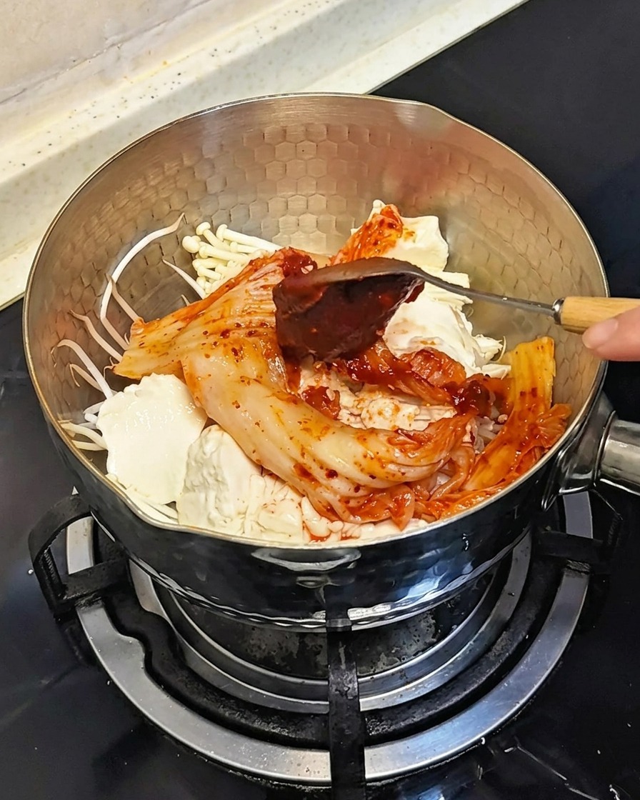 Una cuchara añadiendo una cucharada de pasta de chile coreano roja espesa sobre los ingredientes en la olla.