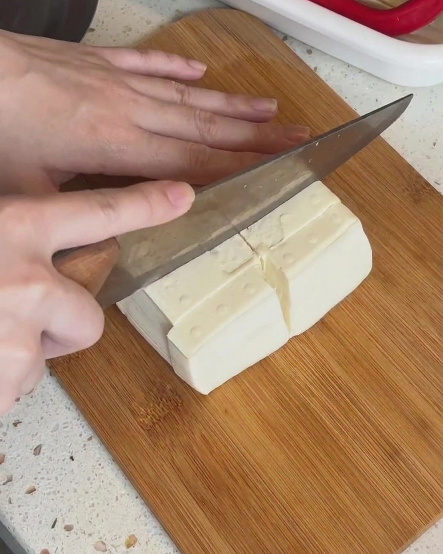 Un bloque de tofu blanco siendo cortado en piezas rectangulares más pequeñas sobre una tabla de madera.