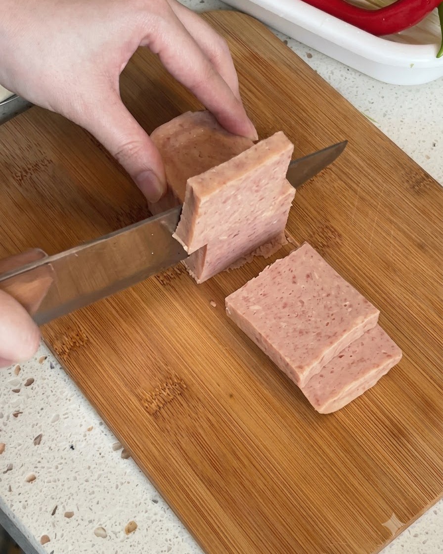 Un bloque de carne en conserva en lata siendo cortado en trozos gruesos sobre una tabla de cortar.