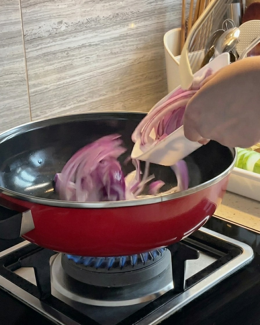 Rodajas de cebolla roja siendo salteadas en una olla roja sobre una hornalla de gas.