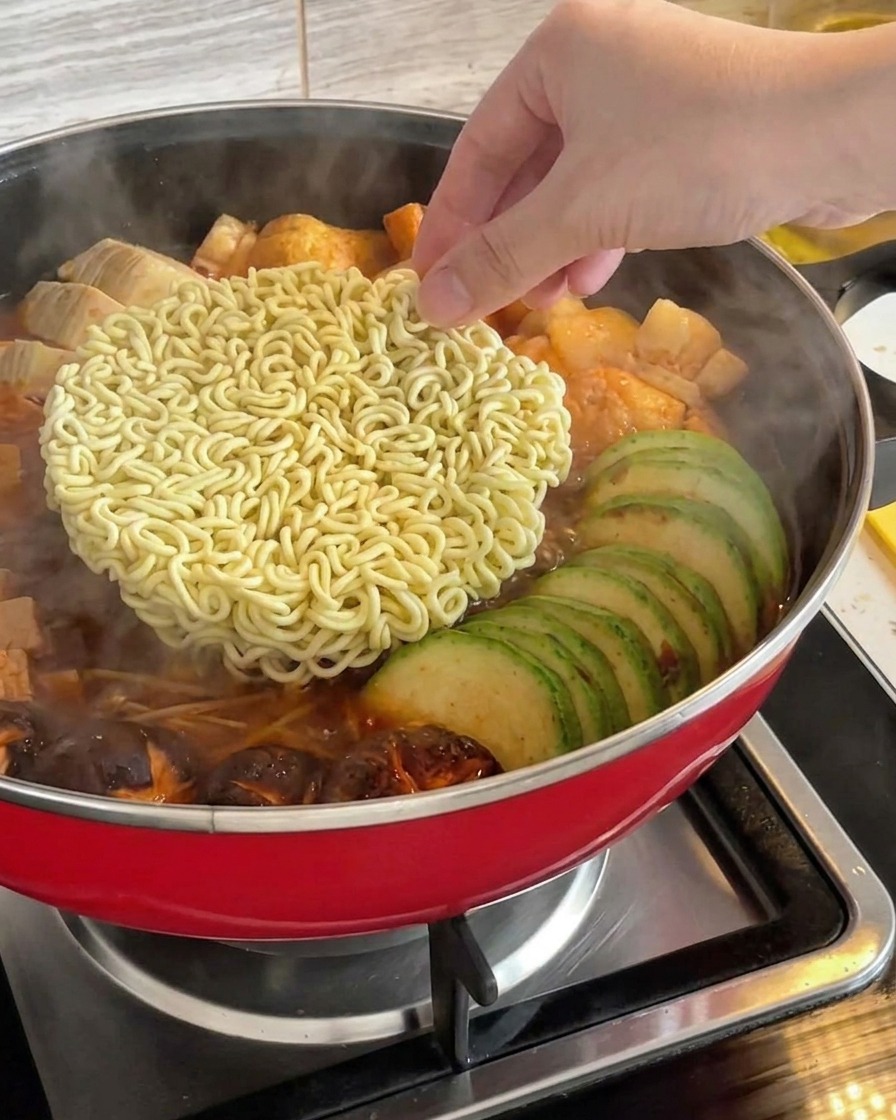 Una mano colocando un bloque de fideos ramen instantáneos secos en el centro de un estofado militar coreano hirviendo a fuego lento.