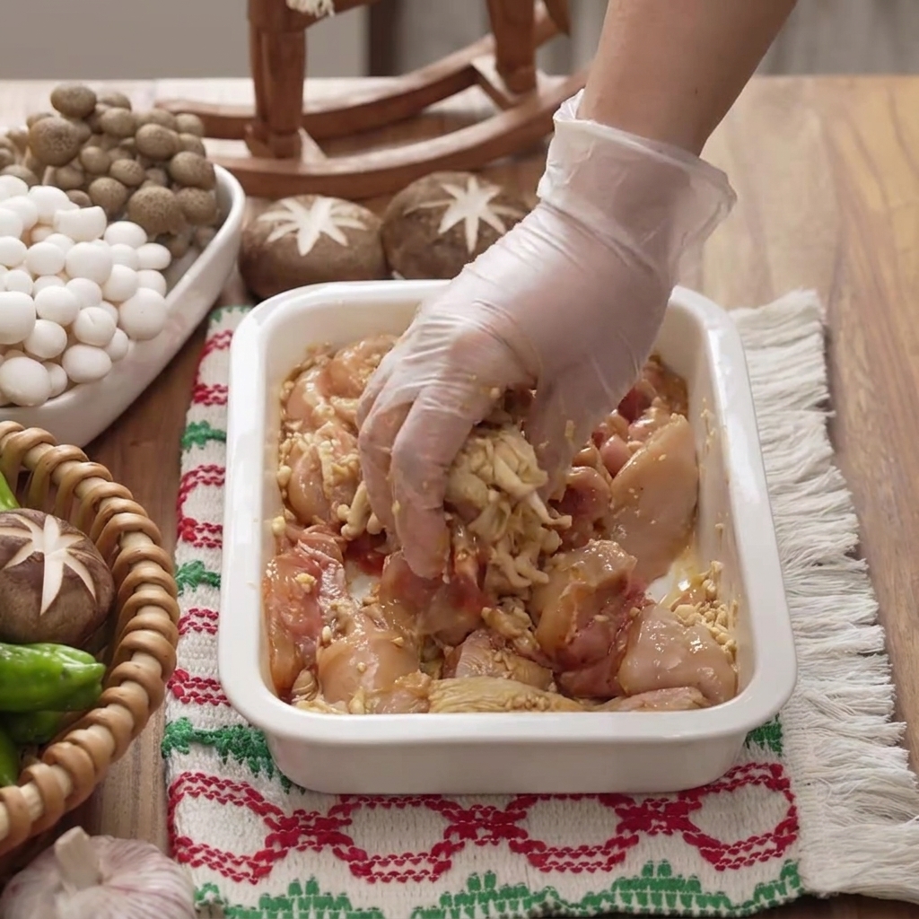 Una mano con guante mezclando y masajeando el adobo en los trozos de pollo dentro de un plato blanco.