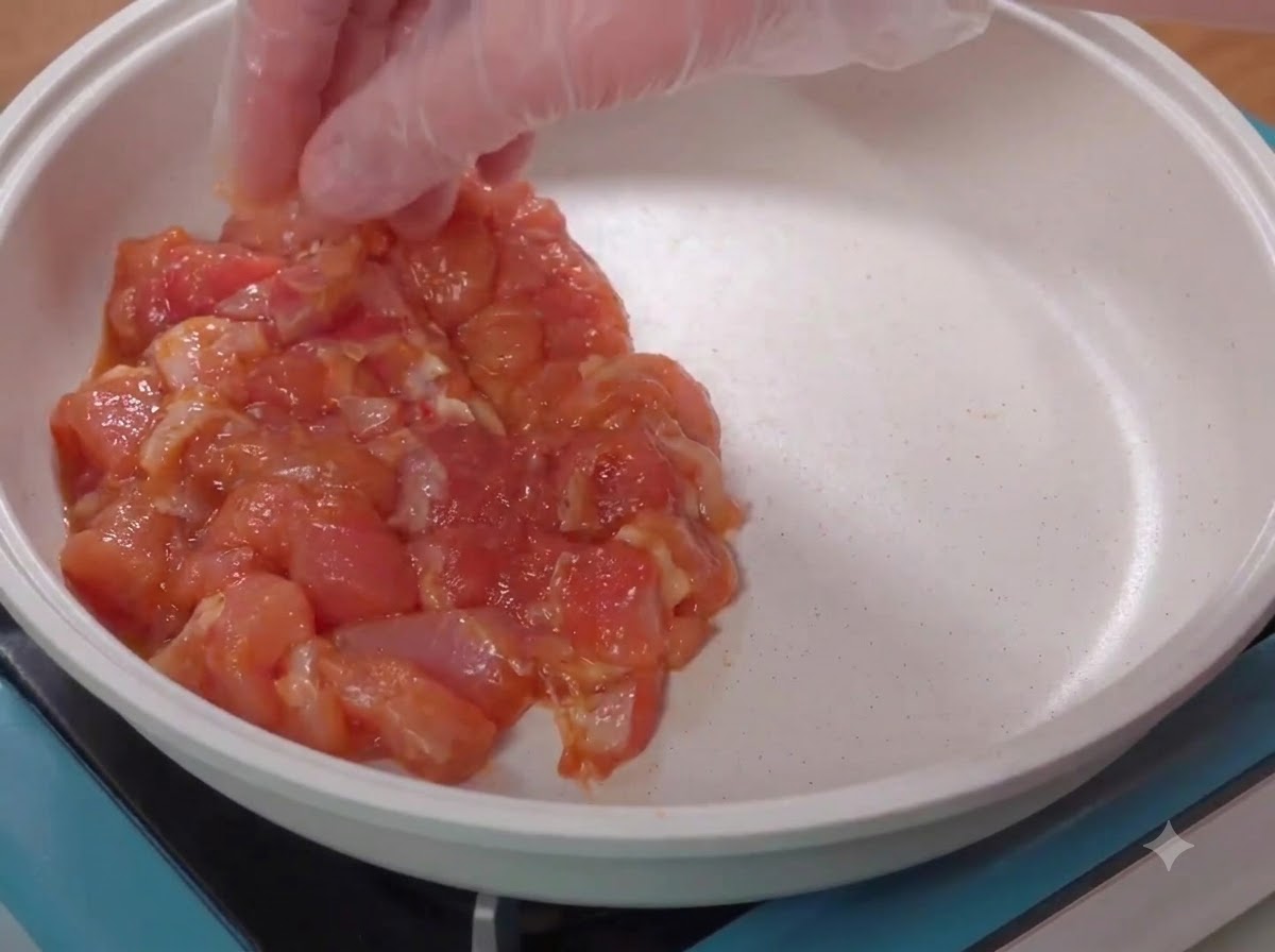 Una mano enguantada colocando trozos de pollo crudo marinados en una sartén blanca para cocinar.