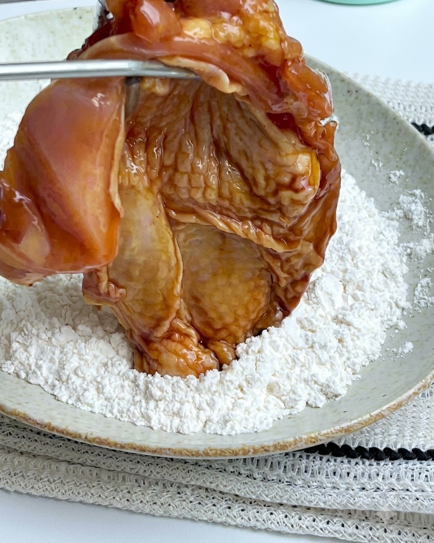 Pinzas sosteniendo una pieza de pollo marinada sobre un plato blanco lleno de polvo de almidón de maíz blanco.