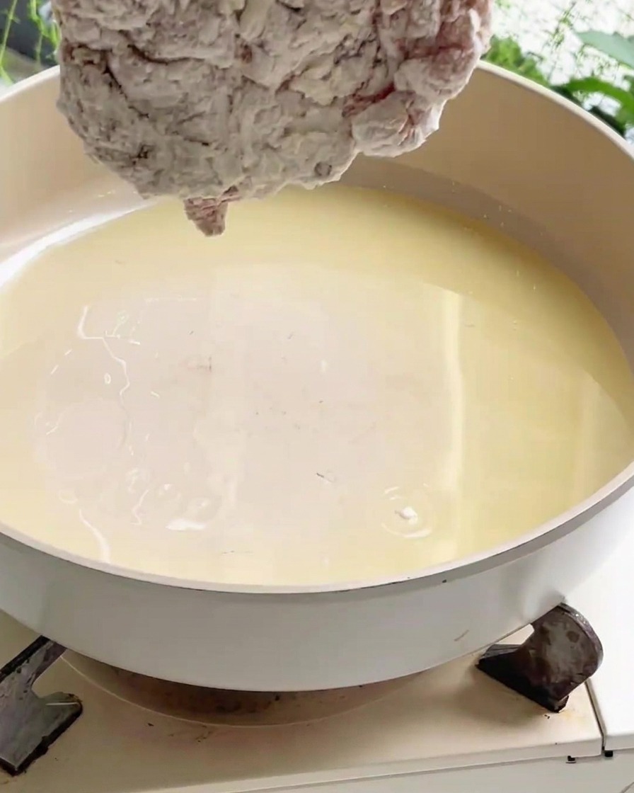 Un trozo de pollo rebozado siendo bajado a una sartén ancha llena de aceite de cocina caliente en una estufa.