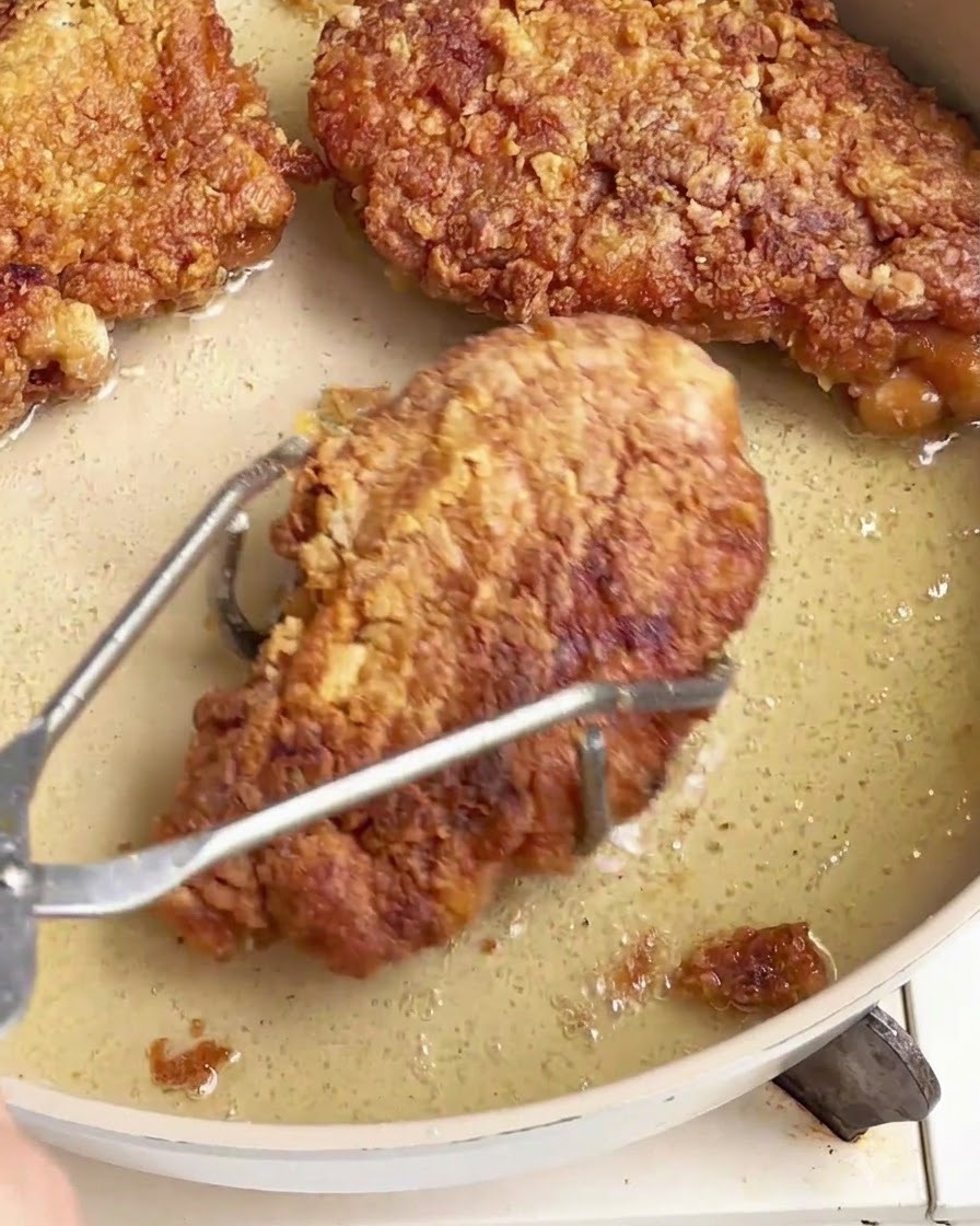 Chuletas de pollo fritas y crujientes siendo retiradas de una sartén aceitosa usando pinzas de acero inoxidable.