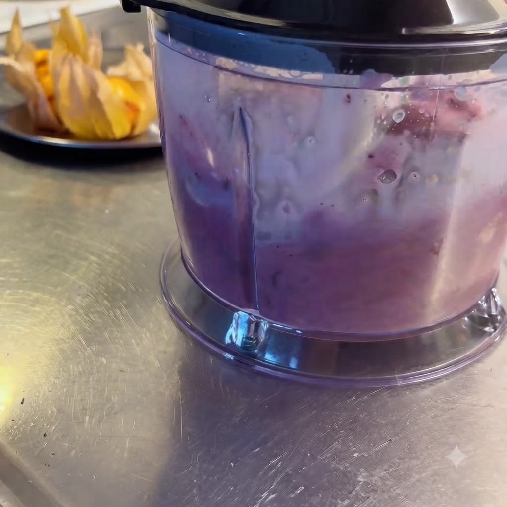 Una licuadora procesando activamente bayas mixtas, frutas y leche hasta formar un batido morado suave.