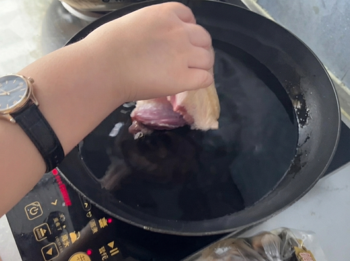Una mano colocando un trozo de cerdo en una sartén con agua fría, jengibre y vino para cocinar.