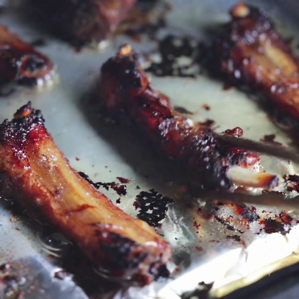 Primer plano de costillas de cerdo a la parrilla siendo volteadas en una bandeja para hornear forrada con papel aluminio usando palillos.