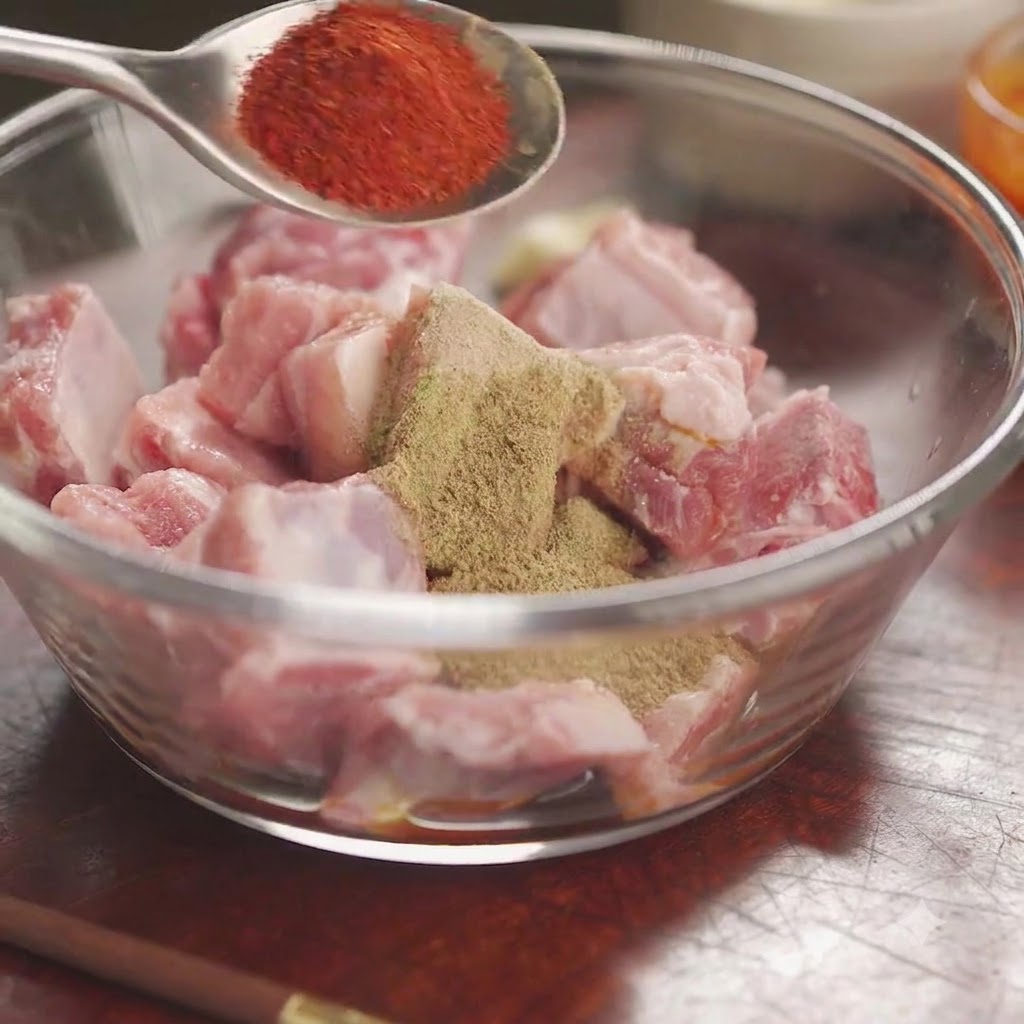 Chile en polvo rojo en una cuchara sostenida sobre un bol de costillas de cerdo crudas cubiertas con especias mixtas y hierbas.