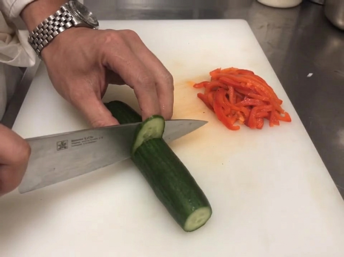 Un cuchillo cortando un pepino verde fresco junto a pimientos rojos en rodajas.
