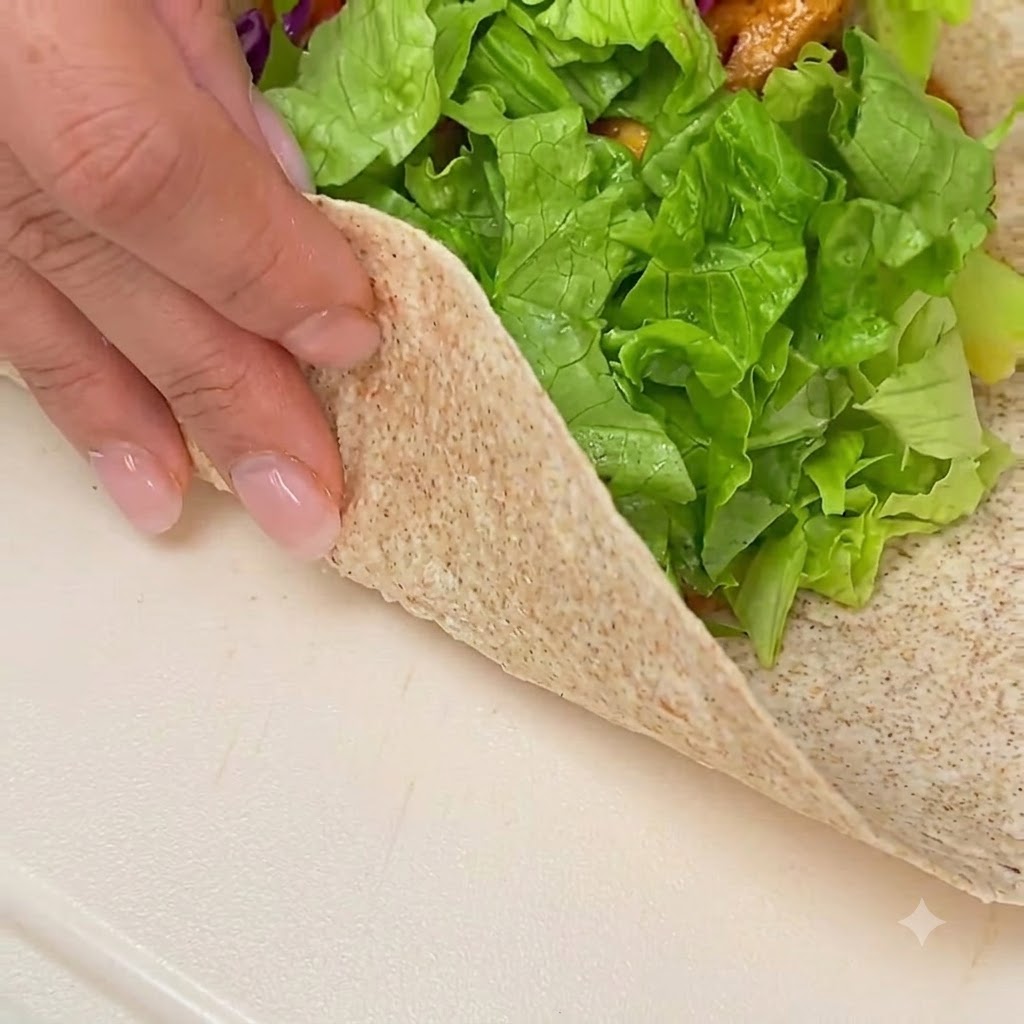 Manos doblando y enrollando una tortilla integral rellena sobre una superficie blanca limpia.