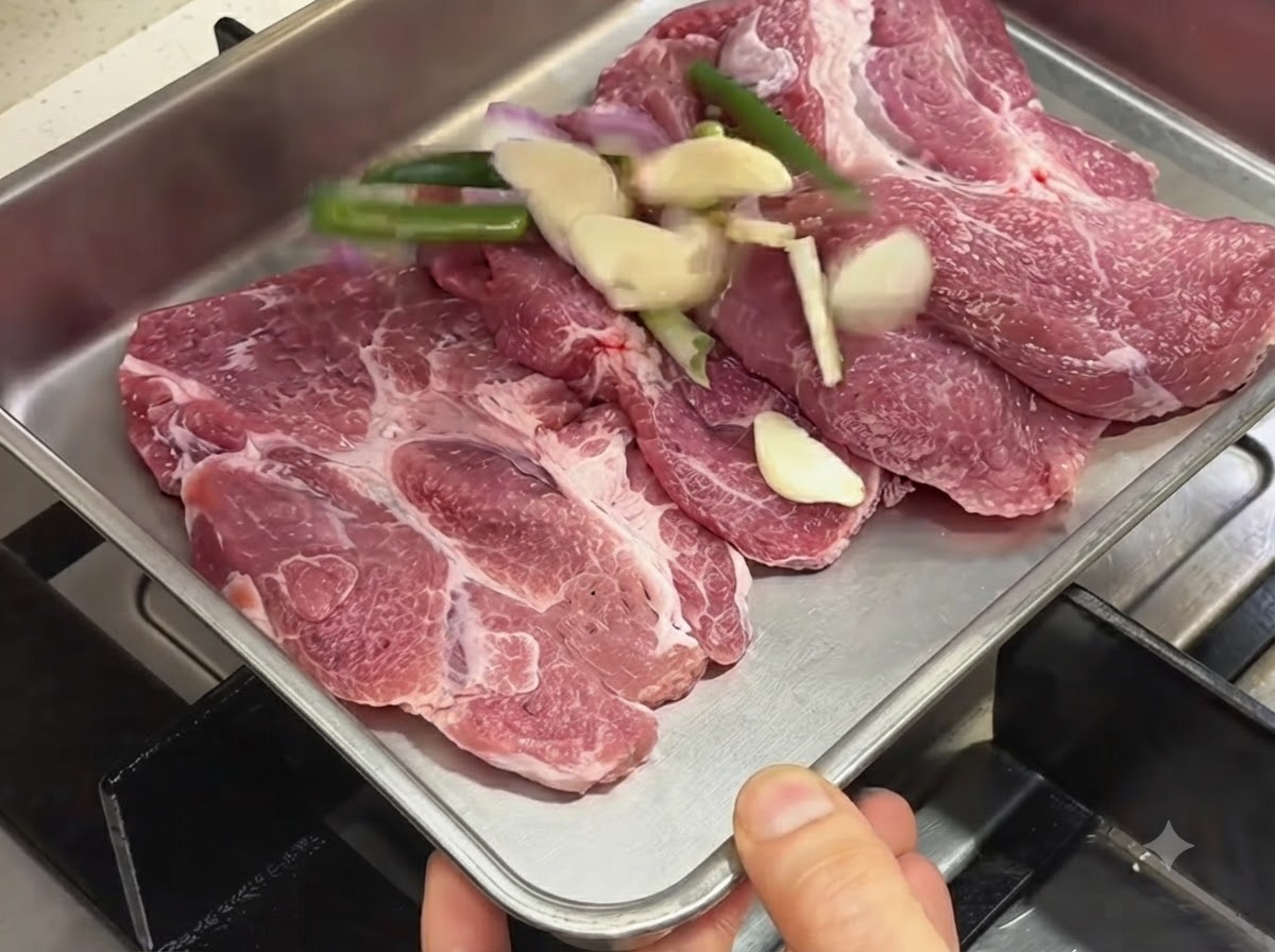 Chuletas de cerdo crudas dispuestas en una bandeja de metal junto a ajo cortado, cebollinos y chalotas.