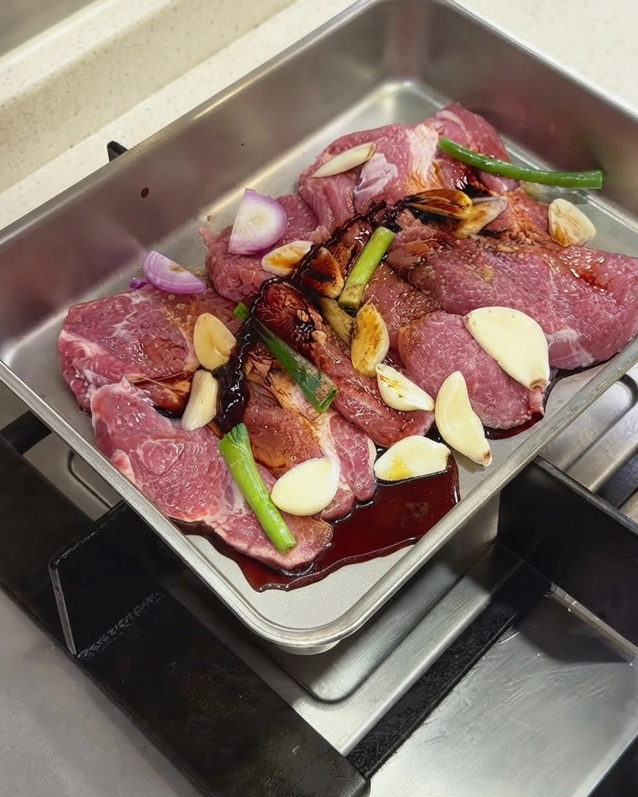 Salsas oscuras y condimentos siendo vertidos sobre las chuletas de cerdo y los aromáticos dentro de una bandeja de metal.