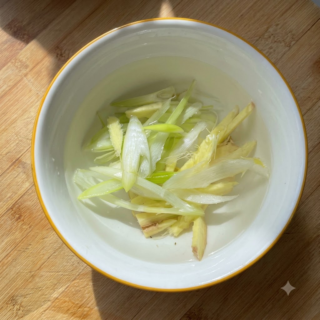 Cebollas verdes picadas y trozos de jengibre macerándose en agua dentro de un pequeño bol blanco.
