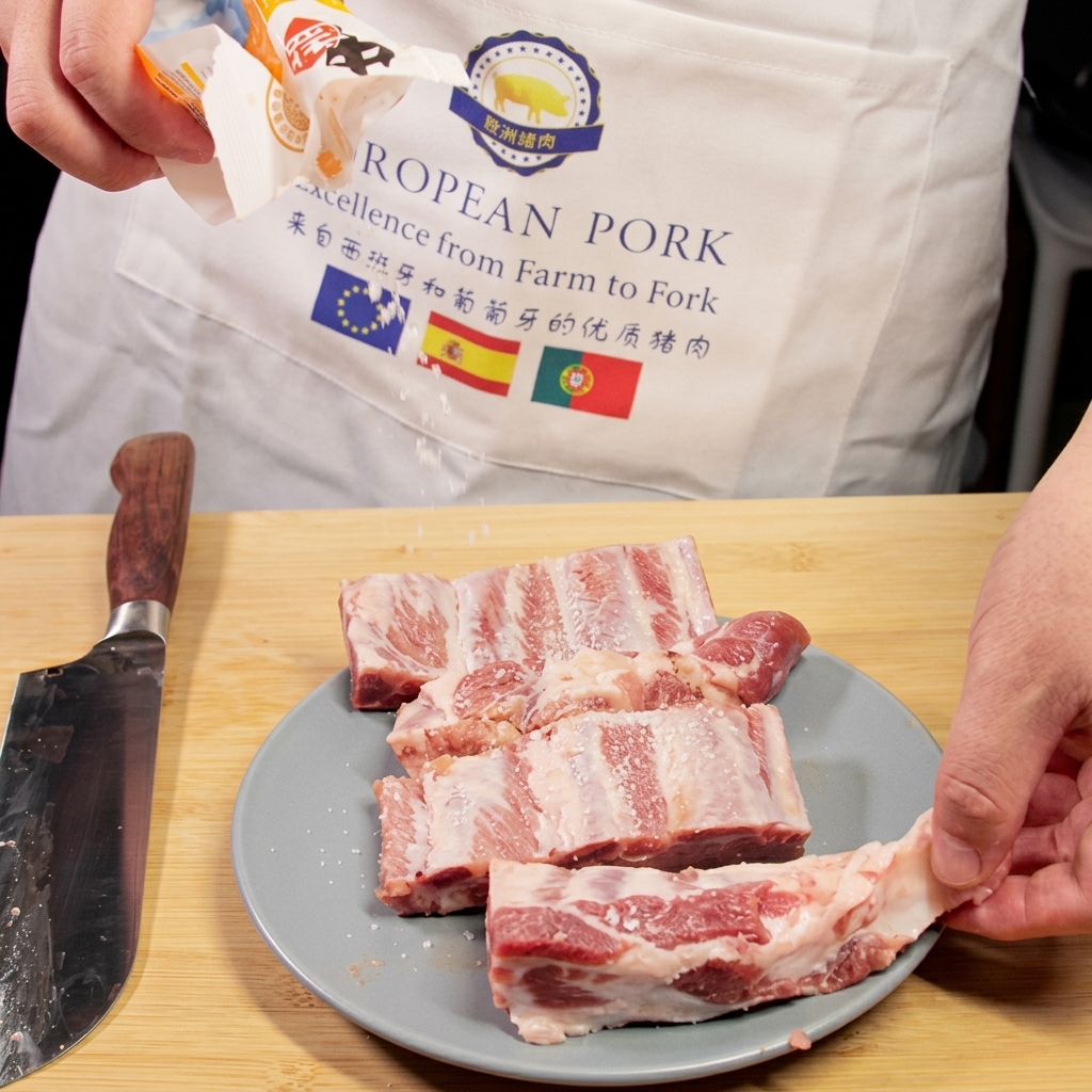 Sal siendo espolvoreada desde un paquete sobre costillas de cerdo crudas que descansan en un plato junto a un cuchillo de chef.