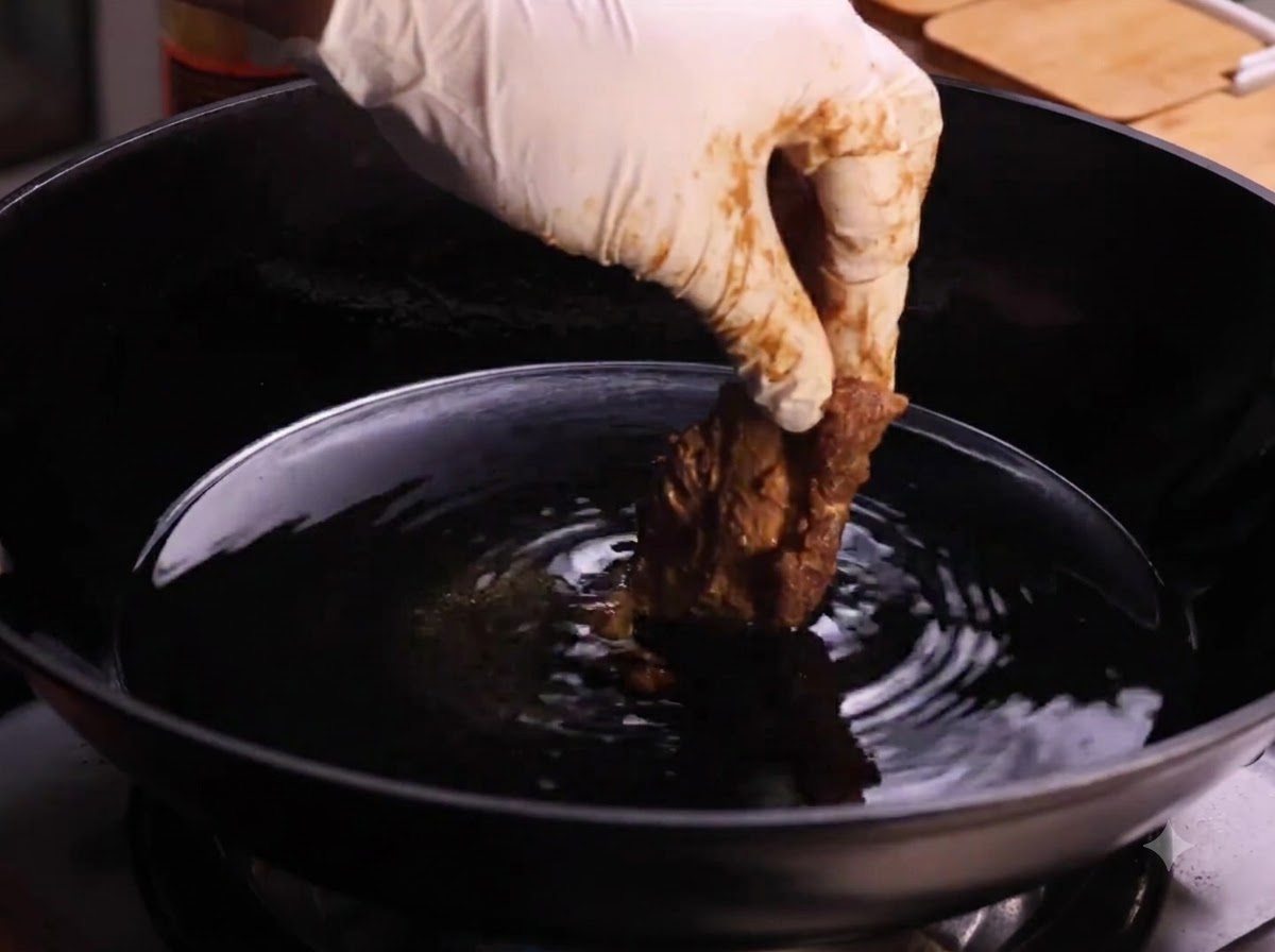 Una mano con guante introduciendo un trozo de costilla de cerdo rebozada en un wok lleno de aceite caliente para cocinar.