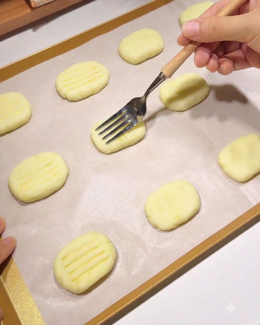 Un tenedor con mango de madera presionando un patrón de líneas paralelas en una tortita de patata moldeada sobre una bandeja para hornear forrada con papel pergamino.